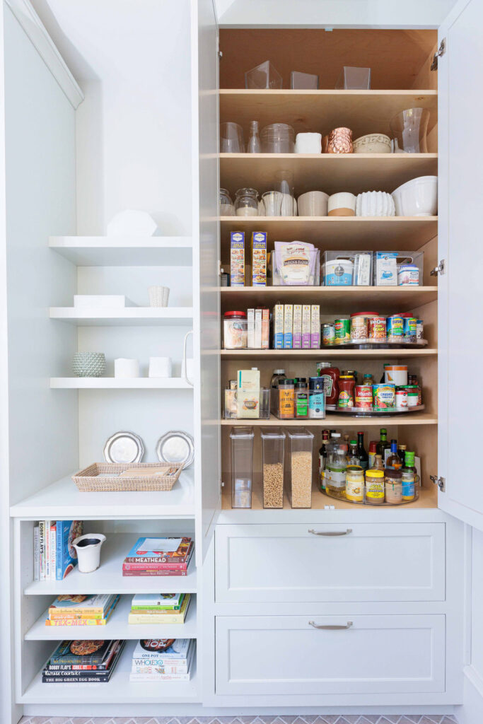 A perfectly organized pantry. The cabinets are white with silver handles, and the inside is a warm wood tone. The contents of the pantry are organzied on turntables, in clear acrylic bins, and in clear acrylic cereal decanters. To the left of the pantry there are stacks of cookbooks, and open-concept shelves holding various pieces of china.