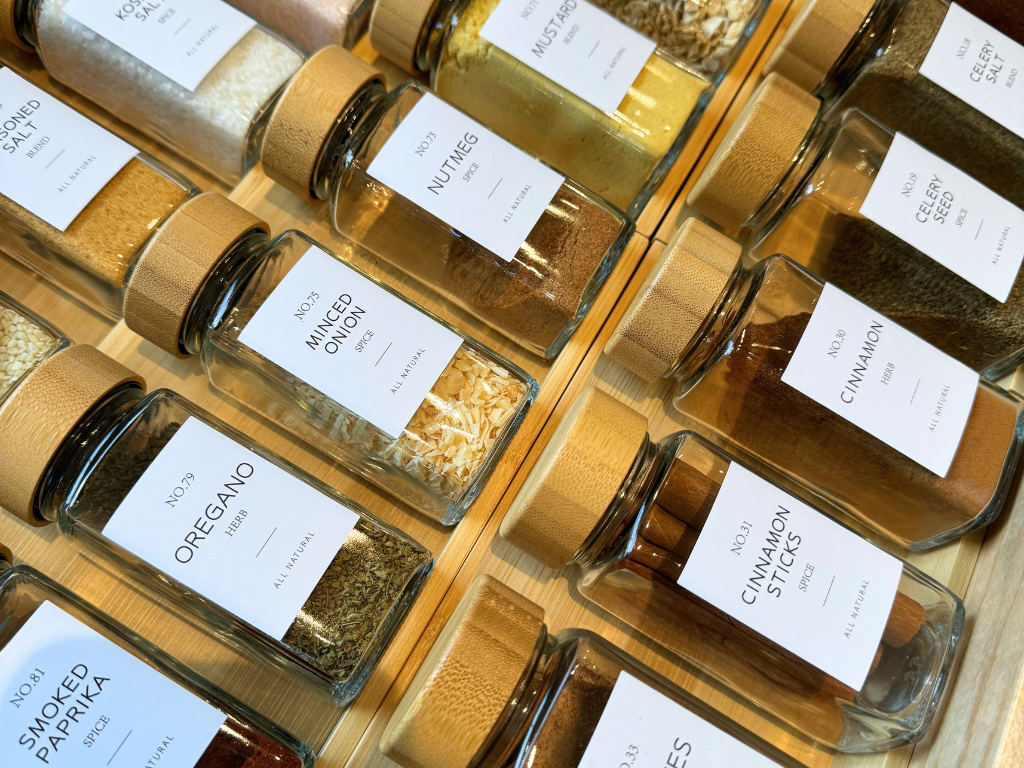 Photo of a variety of spices in glass jars with white labels with black letters. Perfectly organized in a bamboo drawer divider. 