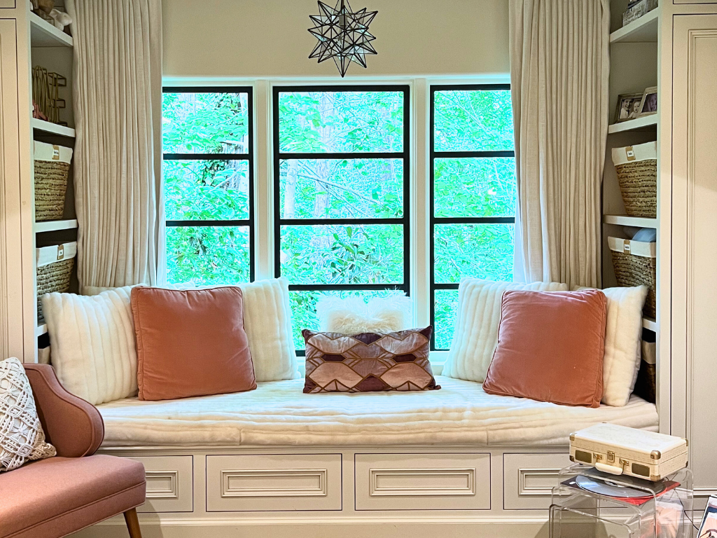 A reading nook daybed in front of a large window with soft white and rose colored bedding